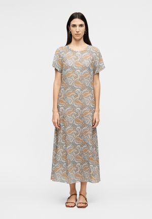 Femme debout portant une robe midi à manches courtes à motif cachemire beige et bleu, avec des sandales marron sur un fond uni.