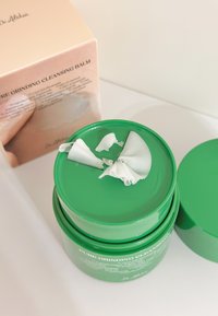 Zielony balsam oczyszczający w okrągłym pojemniku z gładką pokrywką. Kawałek miękkiego, białego balsamu lekko wystaje. Pudełko pokazuje nazwę produktu i szczegóły.