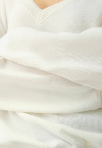 Biały sweter wykonany na drutach z dekoltem w serek. Materiał jest miękki, o delikatnej prążkowanej fakturze, z łagodnymi fałdami i układającymi się marszczeniami.