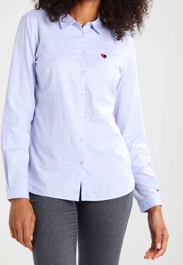 Femme portant une chemise bleu clair à boutons avec un petit logo en forme de cœur sur la poitrine et un pantalon gris foncé, debout devant un fond uni.