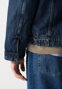 Jeansjacka i mörkblå färg med framträdande sömmar, knäppknapp på ärmslut och ribbat beige nederkant. Strukturerad tyg synlig.