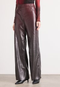 Wijd-valige bordeauxrode leren broek met een gladde textuur, elastische tailleband en subtiele stiksels. In combinatie met zwarte puntige hakken.