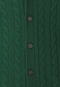 Grüne Strickjacke mit Kabelmuster, die sechs schwarze Knöpfe auf der Vorderseite hat. Weiche Textur mit gerippten Details.