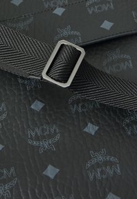 Zwarte leren oppervlakte met een subtiel MCM-logo patroon. Inclusief een gestructureerde zwarte band met een vierkante metalen gesp.