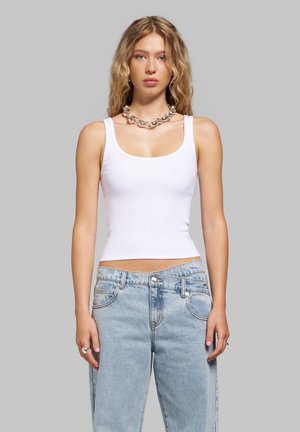 Jeune femme aux cheveux blonds bouclés portant un débardeur blanc, un jean bleu clair, un collier en argent massif et des bagues, debout devant un fond gris.