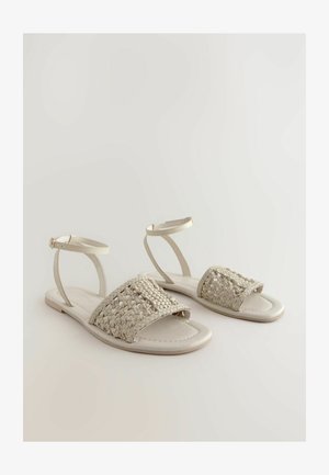 Plochý sandál s tkaným béžovým zvrškom, hladkou svetlou podrážkou a popruhom okolo členka pre podporu. Jednoduchý a otvorený dizajn.