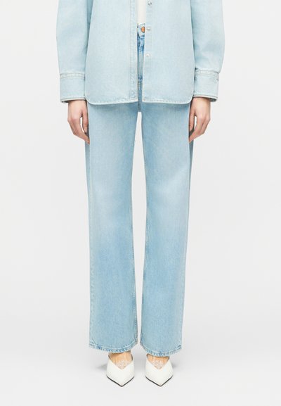 Partie inférieure d'une personne portant un jean large en denim bleu clair, une chemise en denim assortie et des talons blancs à bout pointu avec un détail en dentelle.