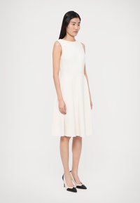 Λευκό αμάνικο φόρεμα με στενή γραμμή στο μπούστο και φούστα που ανοίγει προς τα κάτω. Το μοντέλο φοράει ασπρόμαυρες γόβες με μυτερή μύτη. Απλή και λεία υφή υφάσματος.