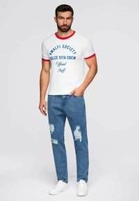 Camiseta blanca con ribete rojo, texto gráfico azul. Combinada con jeans azul desgastados y zapatillas blancas. Ajuste casual, diseño de cuello redondo.