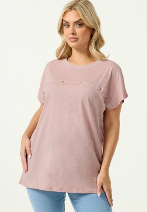 Blondynka w luźnej, krótkim rękawem różowej bluzce z złotymi guzikami na wysokości klatki piersiowej oraz w jasnoniebieskich dżinsach, stojąca na jednolitym tle.