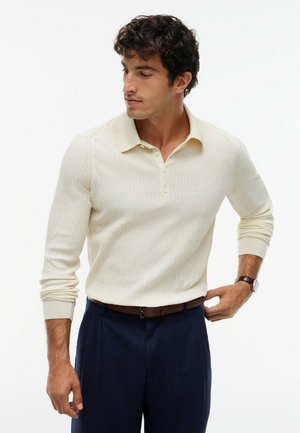 Hombre que lleva una camisa polo de manga larga de canalé color crema metida en pantalones azul marino con un cinturón de cuero marrón y un reloj.