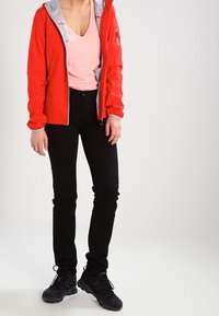 Chaqueta roja con cremallera y forro gris, camiseta de manga corta rosa claro con escote en V, pantalones negros ajustados y zapatillas deportivas negras. Ropa casual para exteriores.