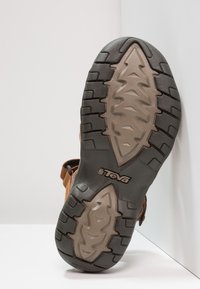 Sandal castanho com uma sola de borracha texturizada e um solado com padrão de folhas, logótipo estampado e correias ajustáveis para um ajuste seguro.