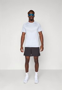 Lys blå atletisk t-skjorte med et teksturert mønster, kombinert med svarte shorts. Modellen har på seg solbriller og hvite joggesko.