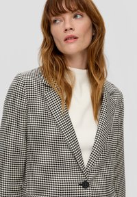 Blazer im Hahnentrittmuster in Schwarz und Weiß, mit schmalem Revers und einer schwarzen Knopfverschluss. Getragen über einem weißen Stricktop.