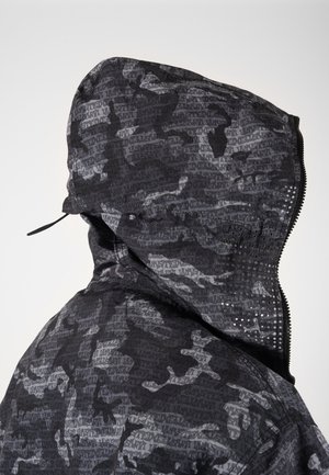 Chaqueta con capucha de camuflaje gris oscuro con cubierta facial con cremallera que presenta pequeños agujeros de ventilación en un lado.
