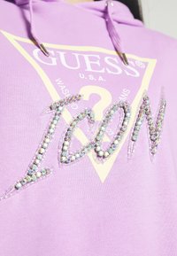 Hoodie di un leggero viola con un logo triangolare giallo con la scritta "GUESS U.S.A.". La parola "ICON" è adornata con strass iridescenti.