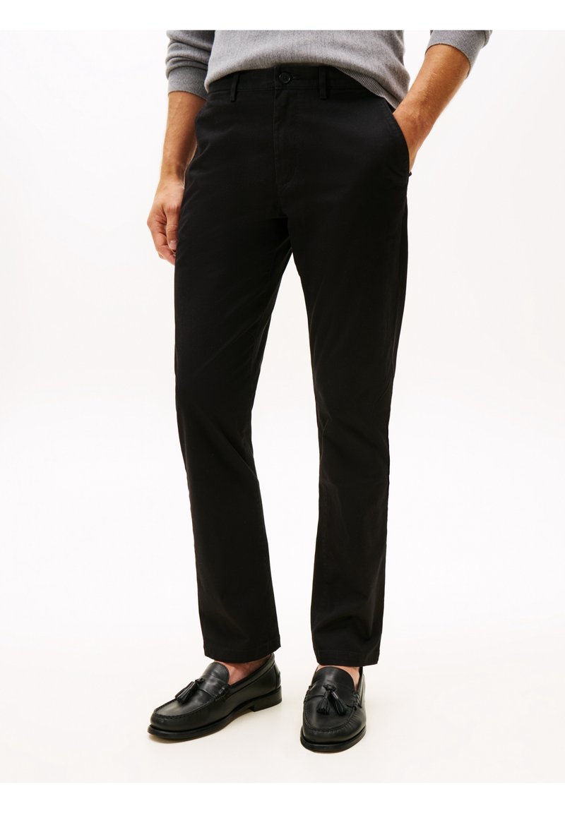 Homme portant un pantalon noir droit, un pull gris et des mocassins noirs en cuir à glands, debout les mains dans les poches.