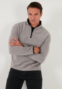 Grå fleece-pullover med kvart lynlås krave, med lange ærmer og sort detaljer ved manchetterne, iført med mørke jeans.