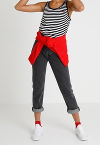 Débardeur à rayures noires et blanches, pull rouge noué à la taille, jean gris foncé retroussé, chaussettes rouges et baskets blanches. Tenue simple et décontractée.
