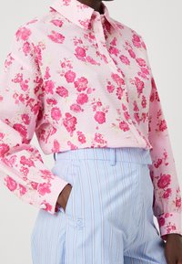 Camicia a maniche lunghe con bottoni di colore rosa e con motivo floreale. Presenta una finitura testurizzata e un colletto a punta. Abbinata a pantaloni a righe blu chiaro.