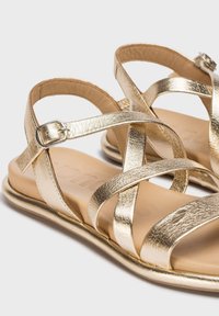 Sandalias metálicas doradas con tiras cruzadas y suela plana. El material tiene un acabado texturizado similar al cuero y un detalle de hebilla seguro.