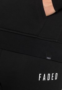 Felpa nera in cotone con tasca a marsupio e polsini a coste. Presenta un piccolo etichetta "FADED" e lettering bianco sul lato inferiore.