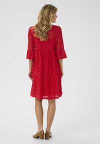 Red lace dress with three-quarter bell sleeves, floral pattern, flared hemline, and paired with beige block-heeled shoes.