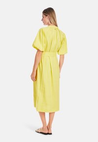 Gelbes Leinenkleid mit kurzen Puffärmeln und einem taillierten Gürtel, das einen geraden Schnitt bis zur Mitte der Wade hat und einen dezenten Rückenschlitz aufweist.