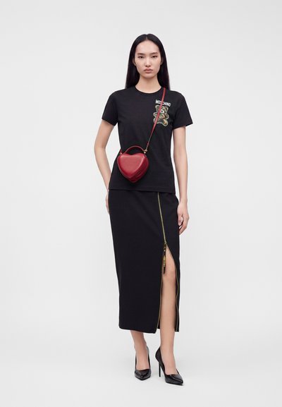 Femme portant un t-shirt noir Moschino avec un ours en peluche, une jupe noire avec une fente zippée à l'avant, des talons noirs et un sac bandoulière rouge en forme de cœur.