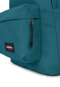 Tealfärgad tygryggsäck med en framficka med dragkedja. Har en svart Eastpak-logotyp och en strukturerad yta. Inkluderar en svart dragsko.