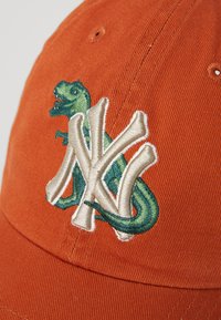 Orange Kappe mit strukturiertem Finish. Besitzt einen gestickten Dinosaurier und das "NY"-Logo in Creme und Grün.