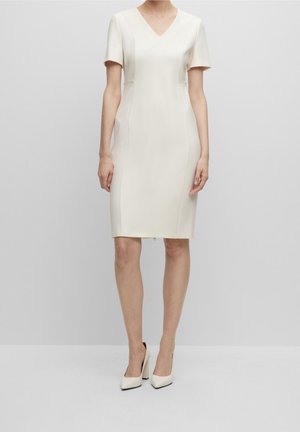 Frau trägt ein knieumspielendes, kurzärmeliges, V-Ausschnitt-Elfenbeinkleid, kombiniert mit passenden elfenbeinfarbenen High Heels, steht vor einem schlichten Hintergrund.