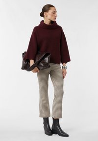 Burgundowa dzianinowa sweter z wysokim kołnierzem, wzorzyste beżowe i brązowe w kratę spodnie oraz czarne botki, trzymając brązową skórzaną torbę.