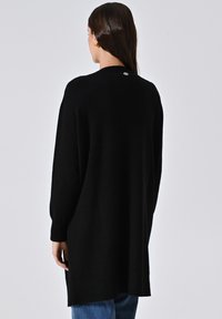 Frau mit langen dunklen Haaren, die einen langen schwarzen Pullover und blaue Jeans trägt, von hinten vor einem einfarbigen Hintergrund gezeigt.