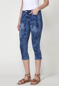 Pantalons capri en denim, bleus avec un motif de feuilles foncées, design ajusté, taille mi-haute et bords retroussés. Associés à des sandales brunes à lanières.