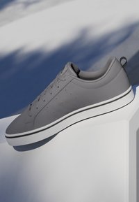 Zapatilla deportiva gris de caña baja con cordones delanteros y suela blanca, exhibida sobre un pedestal blanco, fondo azul sombreado.