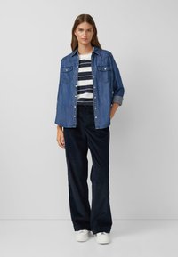 Denimhemd mit spitzem Kragen und Druckknöpfen, getragen über einem gestreiften Baumwoll-T-Shirt. Kombiniert mit dunklen Cordhosen und weißen Sneakers.