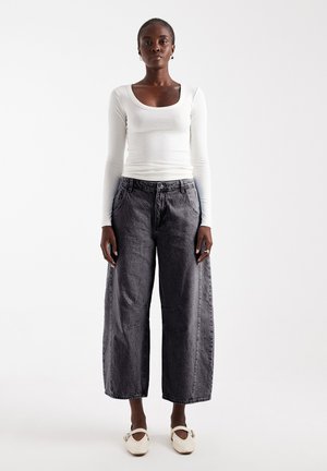 Donna in piedi su sfondo bianco, indossa un top bianco a maniche lunghe, jeans neri sbiaditi a gamba larga, scarpe bianche basse e piccoli orecchini.