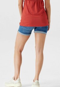 Červený vzorovaný top s volným střihem, spárovaný s ohrnutými džínovými šortkami. Modelka má na sobě světlé tenisky. Jednoduchý, ležérní outfit.