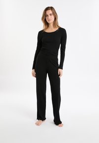 Jonge vrouw die op blote voeten staat in een zwart geribbeld langwerpig shirt en bijpassende wijde broek tegen een vlakke witte achtergrond.