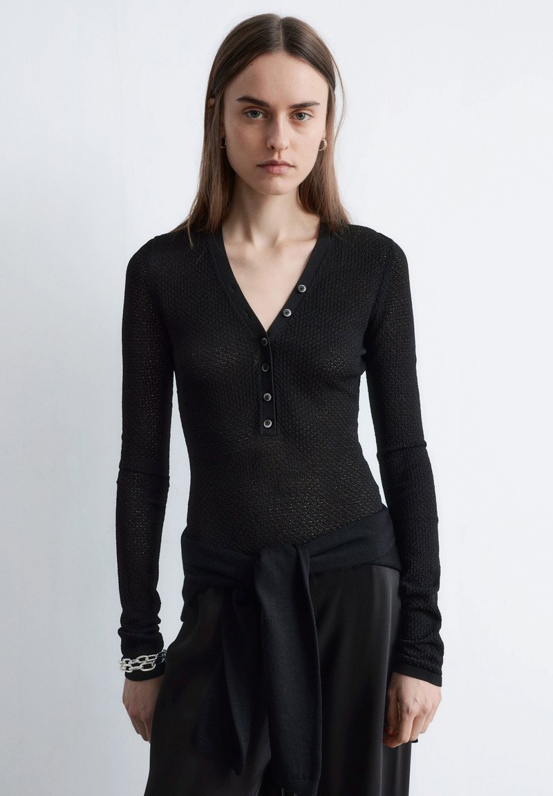 Haut en maille noir à manches longues avec col en V, détail de boutons et tissu texturé, associé à un pantalon noir noué à la taille.