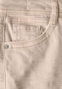 Beige denimtyg med en synlig ficka, silverknappdetalj och kontrasterande orange sömmar längs sömmarna. Sl�ät textur med diagonalt väv.