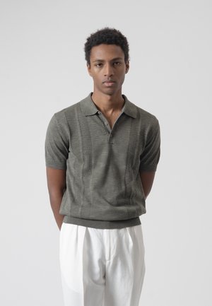 Joven con cabello corto y rizado, vestido con un polo gris de manga corta de punto y pantalones blancos, de pie con las manos detrás de la espalda contra un fondo sencillo.