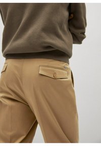 Beige broek met een gladde textuur en twee achterzakken, voorzien van knoopsluitingen en een klein logoaccent. Gepaard met een bruine gebreide trui.