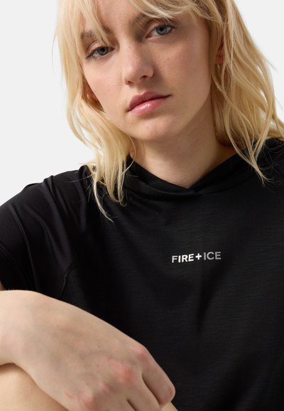 Femme blonde aux yeux clairs portant un t-shirt noir avec le texte "FIRE+ICE", assise avec un bras reposant sur son genou sur un fond blanc.