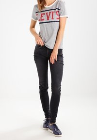 Camiseta de cuello redondo gris con el logo rojo de "LEVI'S", combinada con jeans negros de corte slim y zapatos azules con suelas blancas. Diseño sencillo y textura suave.
