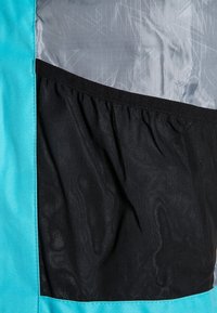 Détail d'une veste turquoise et noire avec une poche en mesh noir extensible et une doublure gris lisse avec un motif diagonal subtil.
