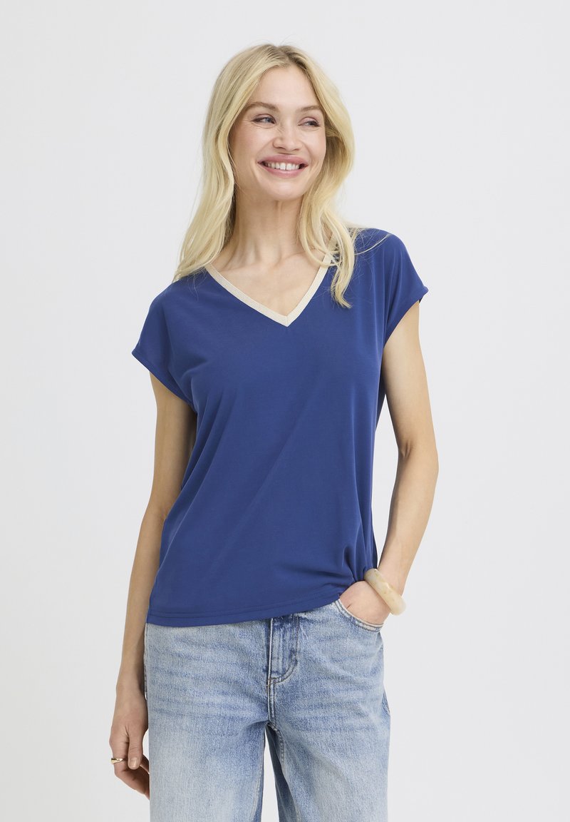 Femme blonde souriante, portant un t-shirt bleu à manches courtes et col en V avec un bord beige, et un jean bleu clair, debout avec une main dans la poche.