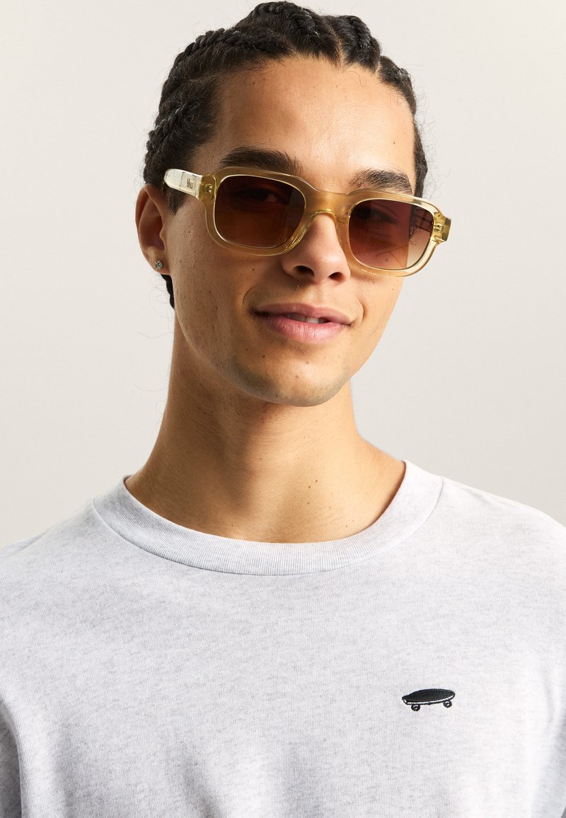Gafas de sol sobredimensionadas con un tono amarillo y un marco transparente. El modelo lleva una camisa gris claro con un pequeño gráfico de patinador en negro.